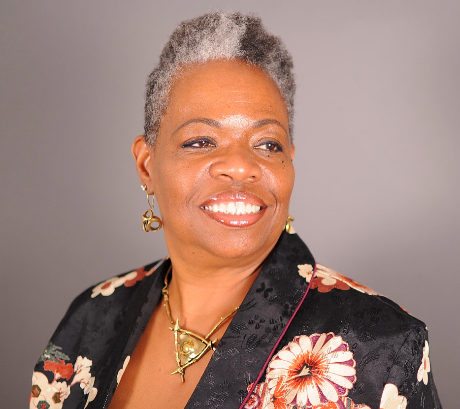 Smiling person with short gray hair wearing gold jewelry and a black floral jacket against a plain background.