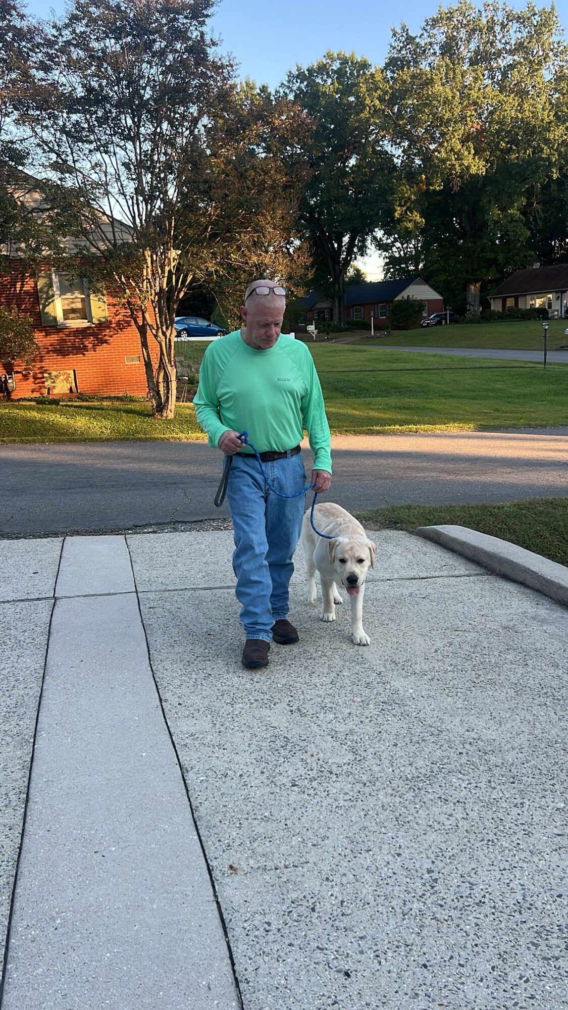 A person stands on a sidewalk beside a small white dog near a park path.
