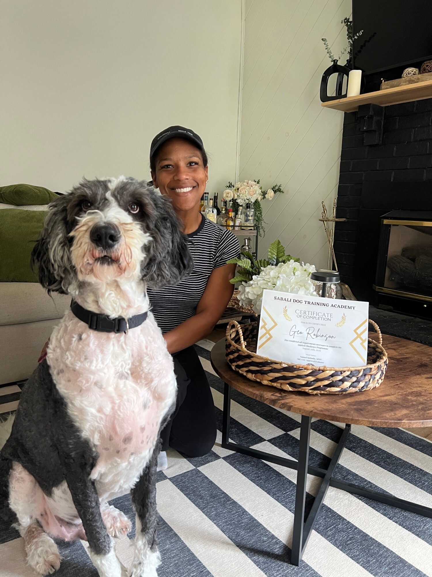 A person sits beside a black-and-white dog, with a woven basket on a chair nearby.
