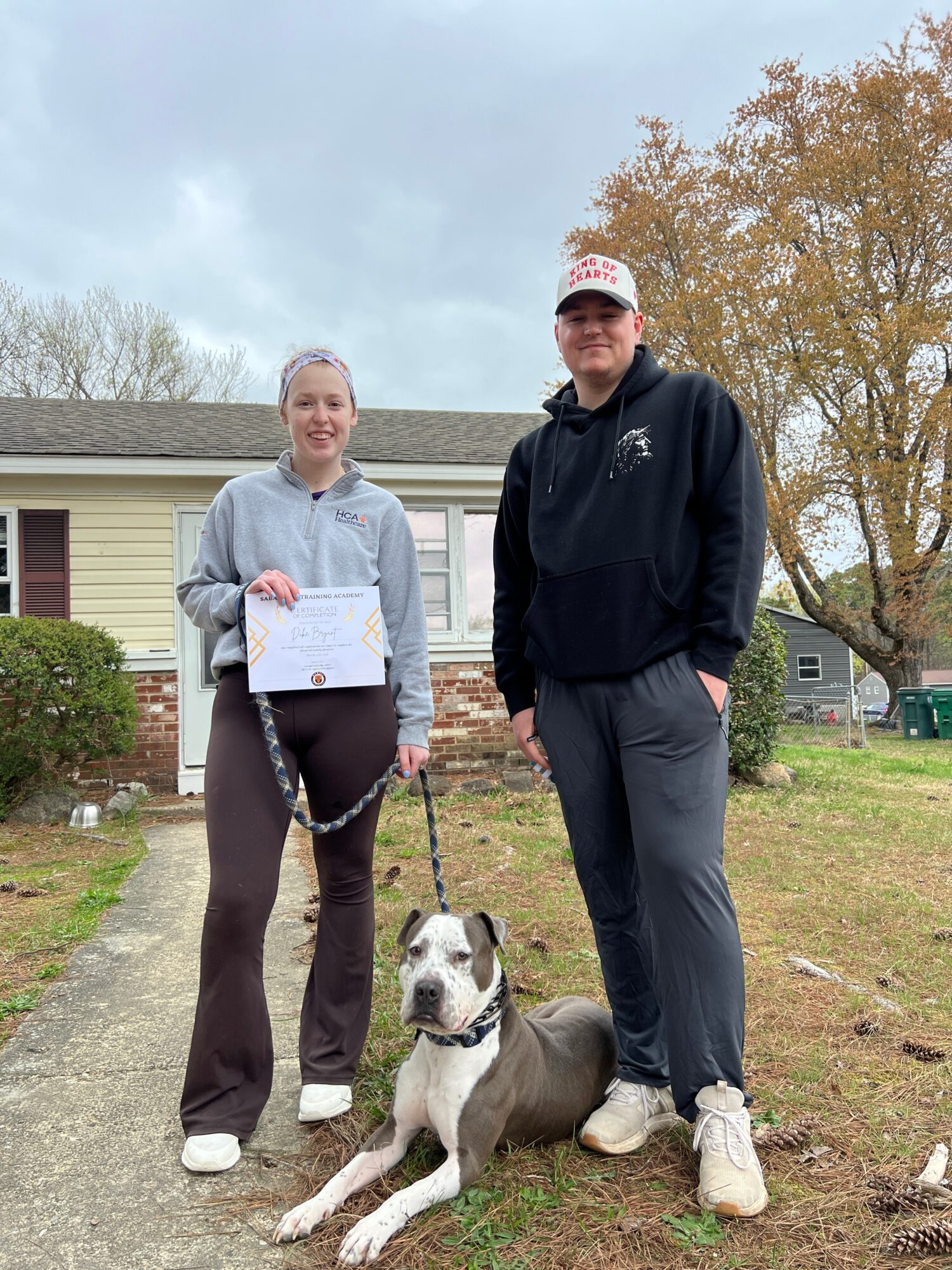 Two people stand on a driveway beside a dog, with a house and trees behind them.