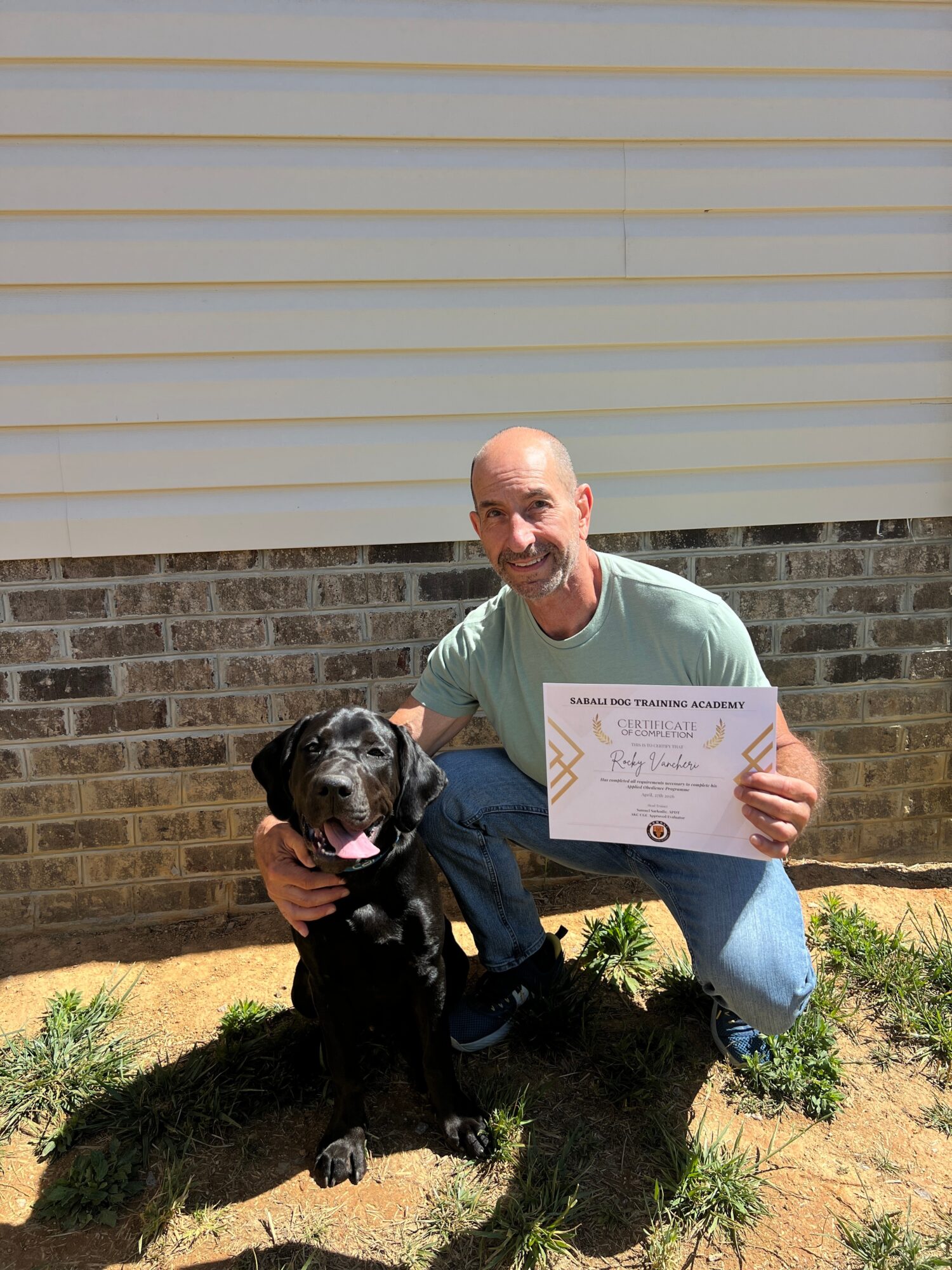 A man kneels beside a black dog, holding a certificate in front of a brick wall and closed window shutters.