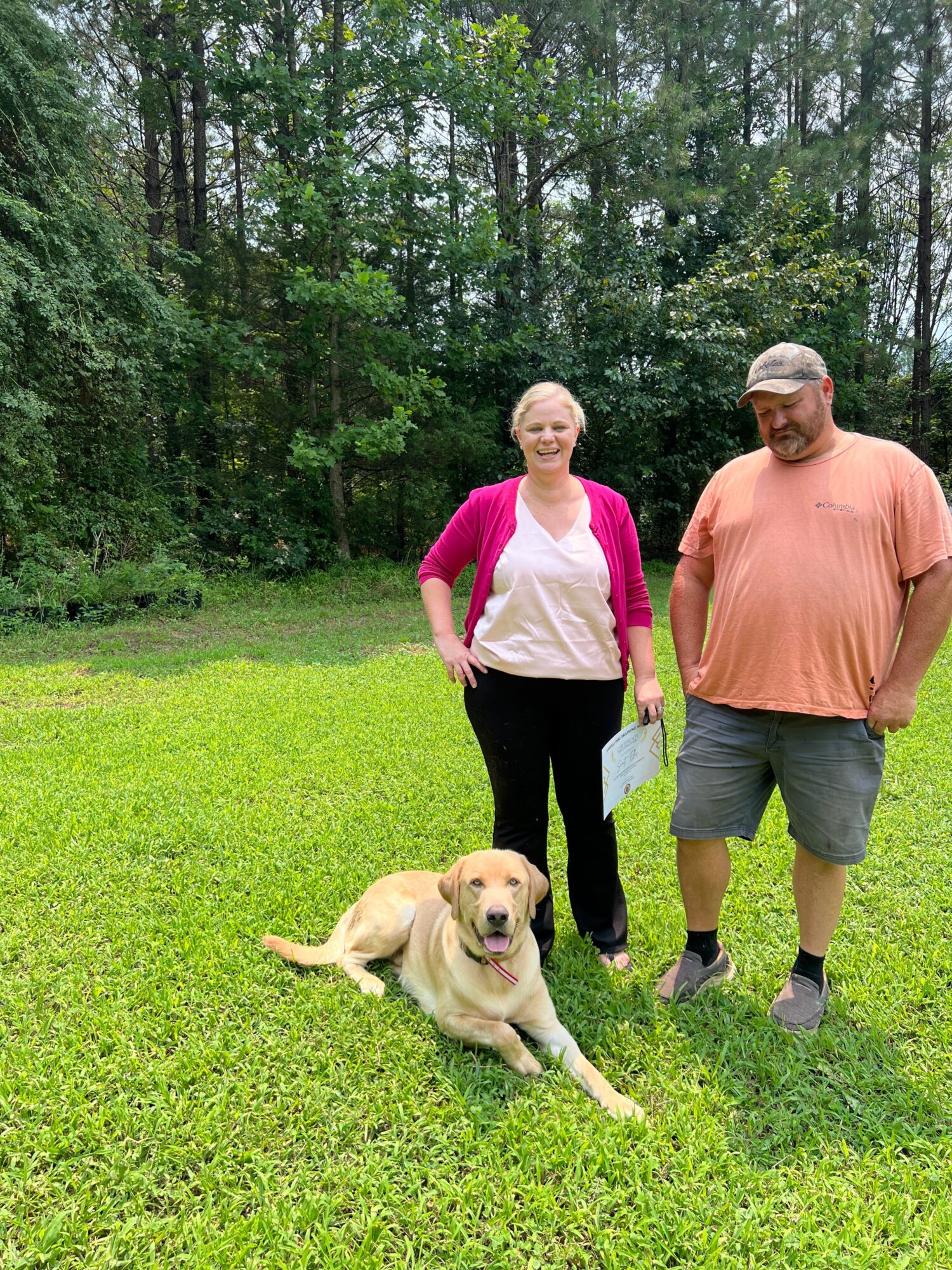 Two people stand on a grassy lawn beside a seated dog, with trees in the background.