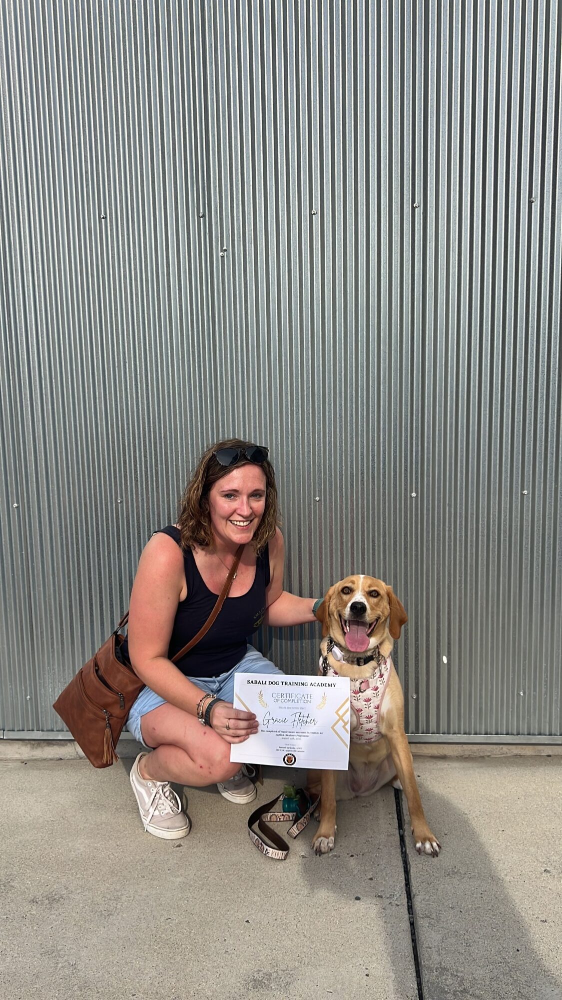 A seated person holds a certificate beside a small dog in front of a metal shutter.