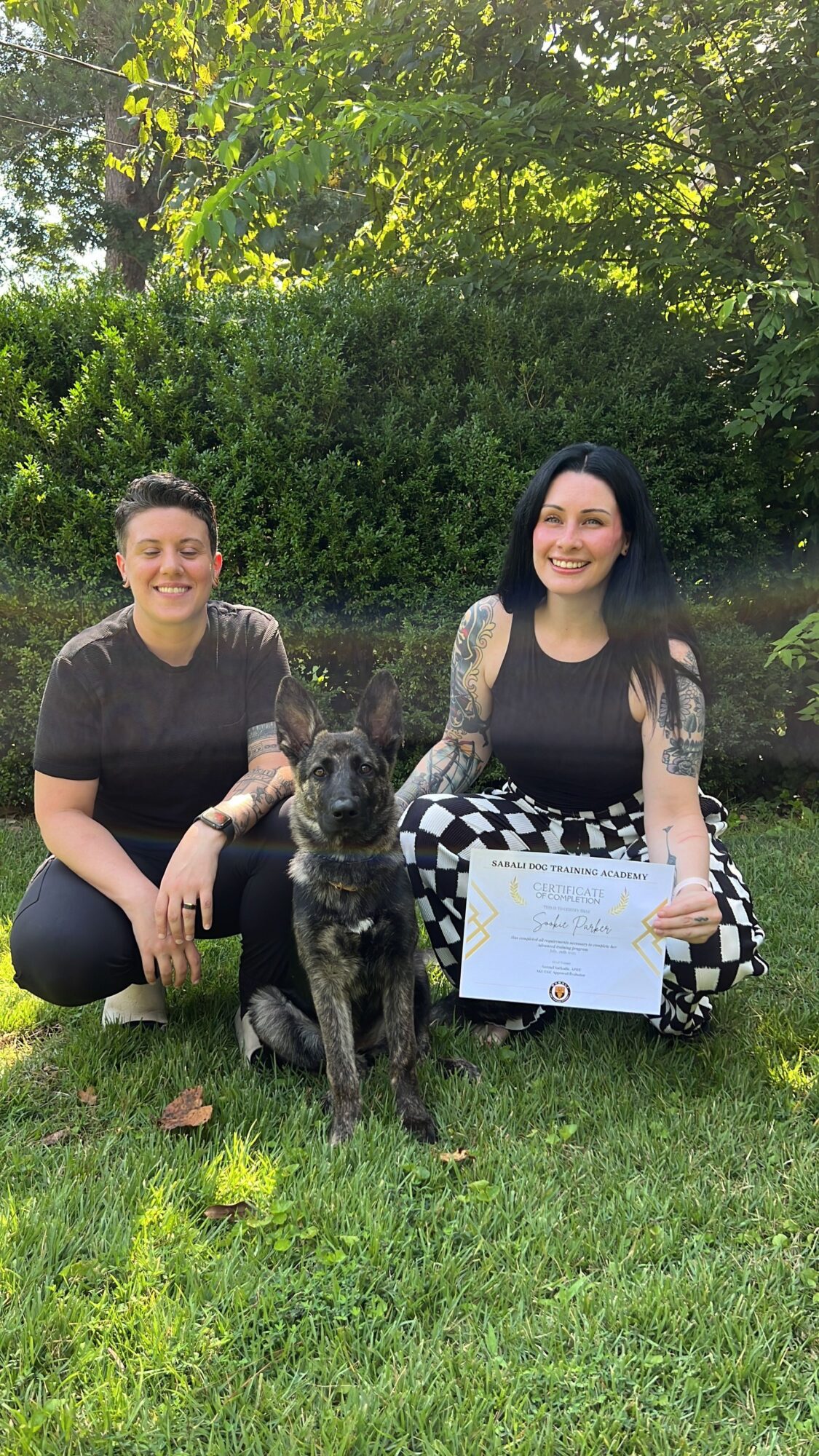 Two people sit on grass near trees, holding a framed certificate.
