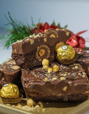 A chocolate dessert topped with sliced chocolate roll, golden wrapped chocolates, hazelnuts, and crumbs on a plate.