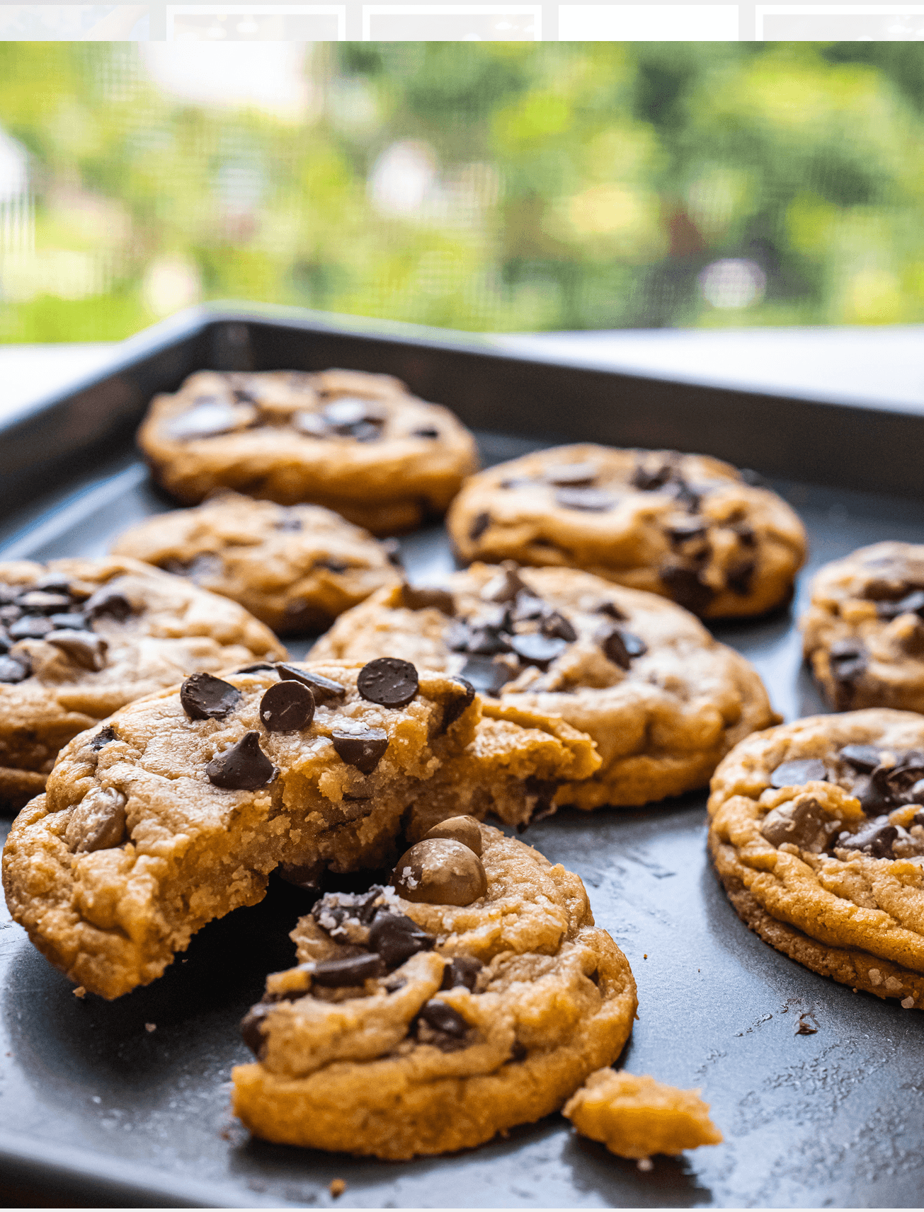 Chocolate chip cookies on a dark baking tray, with a bitten cookie in the foreground.